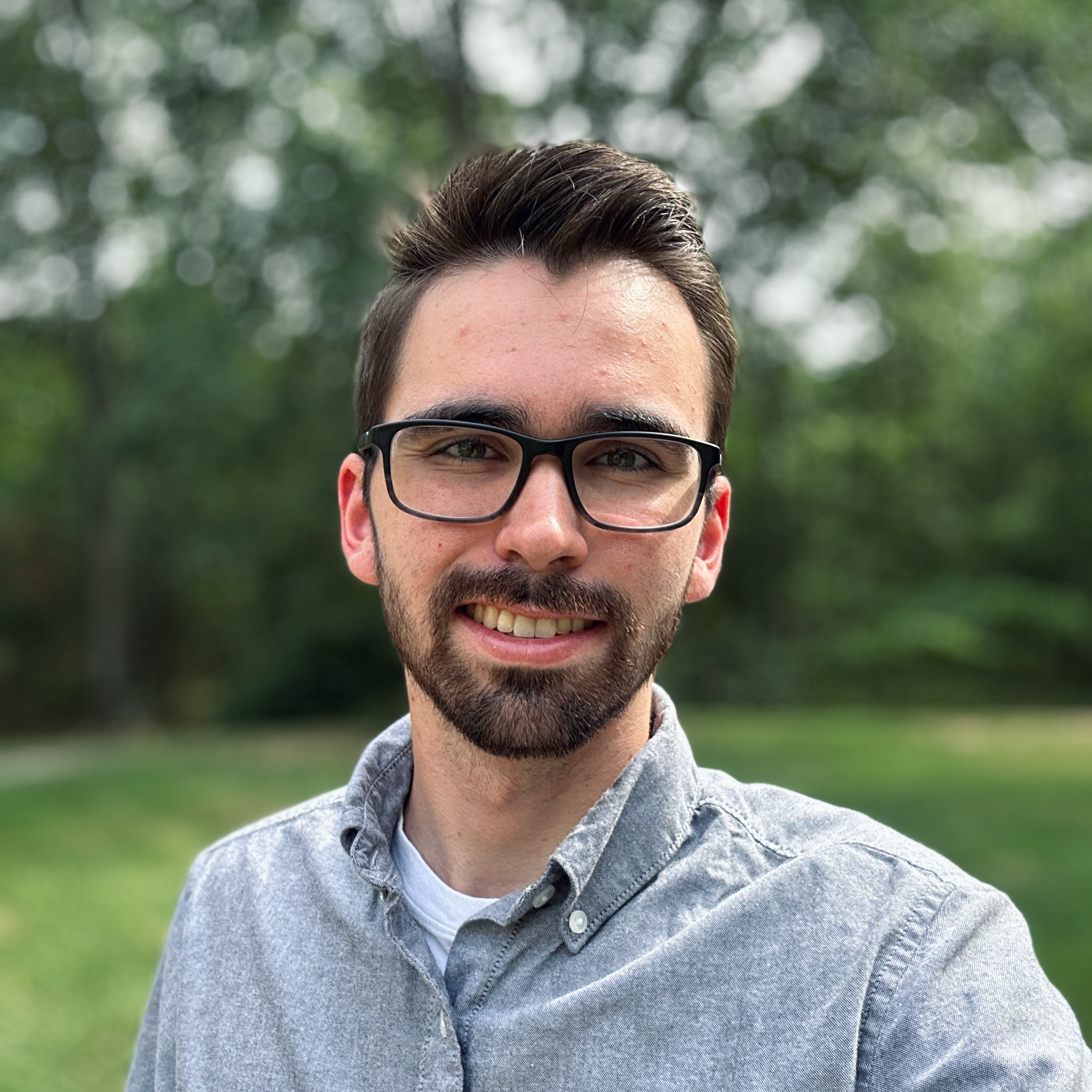 young man with black hair, beard, and glasses wearing a grey button up shirt standing in front of trees